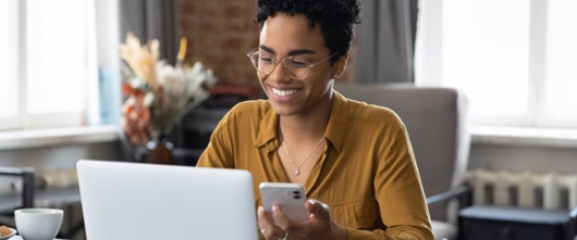 Woman smiling looking at her laptop while also using her phone. 