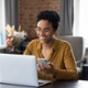 Woman smiling looking at her laptop while also using her phone. 